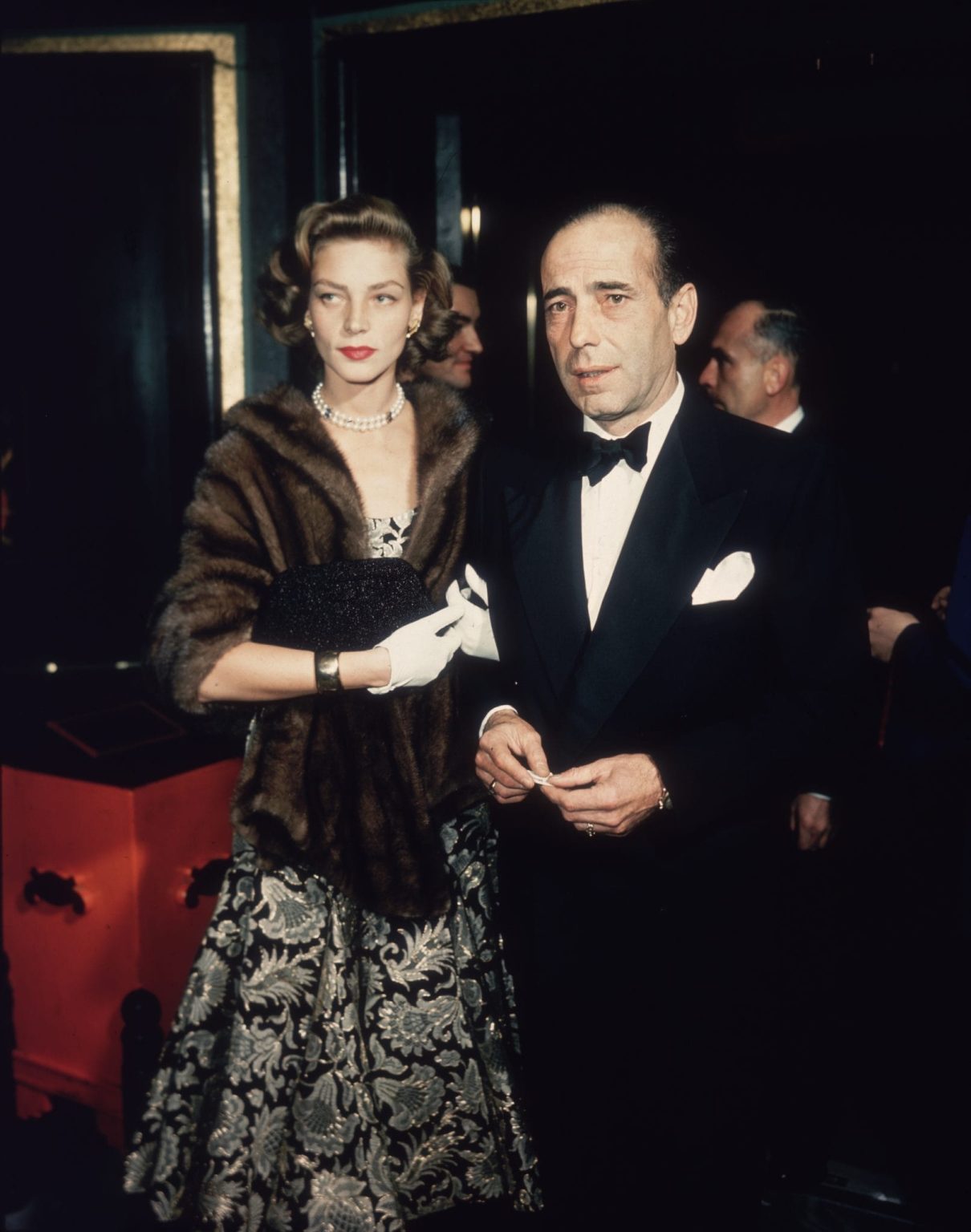 circa 1955: Married American actors Lauren Bacall and Humphrey Bogart (1899-1957) stand in a foyer after arriving together. Bacall wears a brocade dress, a mink wrap, and a pearl necklace. Bogart wears a tuxedo. (Photo by Hulton Archive/Getty Images)